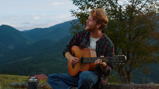 Romantic millennial sing nature in mountains. Close up smiling guy relax on travel camp. Young guy practice music play acoustic guitar outdoors. Relaxed tourist chill on green hill. Leisure concept.
