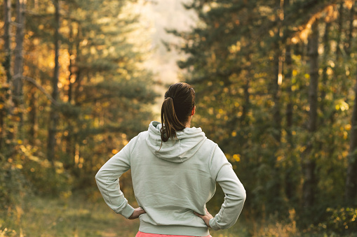 Young female athlete standing in the middle of a forest. Young woman working out in a public park on an early morning. Young woman doing her morning routine, having a walk through the park.
