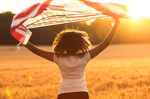 Rear view of mixed race African American girl teenager female young woman in a field holding USA stars and stripes flag above her head in golden sunset evening sunshine