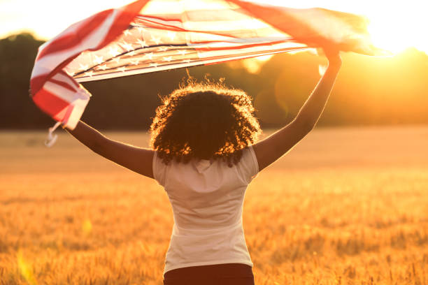 Mixed Race African American Girl Teenager With US Flag at Sunset Rear view of mixed race African American girl teenager female young woman in a field holding USA stars and stripes flag above her head in golden sunset evening sunshine cute teen pic stock pictures, royalty-free photos & images