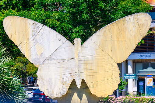 Alicante, Spain - November 5, 2022: La Mariposa (The Butterfly) sculpture by Manolo Valdes. The metallic sculpture covers parts of a tree, and a building is in the background.