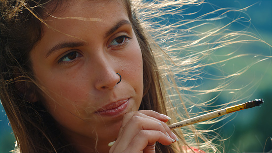 Closeup inspired artist working picture on summer windy day. Focused woman painter touching lips by hands with paintbrush outdoor. Portrait of thoughtful impressionist creating artwork outside.