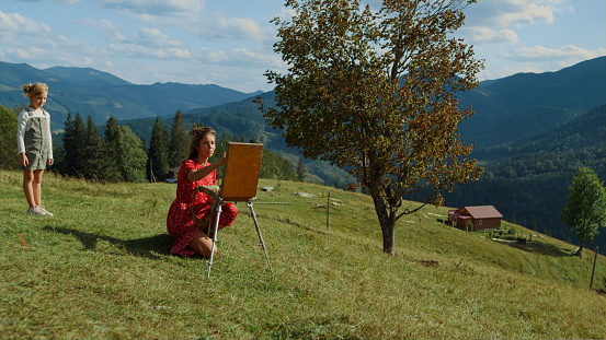 Focused woman painter drawing on easel outdoor. Cute girl watching at creative process on green meadow. Talented woman painting picture mountains in summer vacation sitting grass. Talent concept