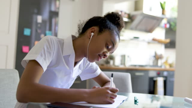 Teenage schoolgirl at home doing her homework while listening to music with earphones