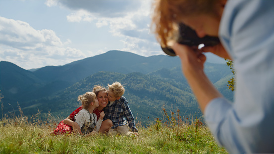 Back view of redhead man photographing smiling woman with kids in front green mountains. Beautiful family enjoying photo session outdoors in summer holiday. Adorable children kissing mother on camera.