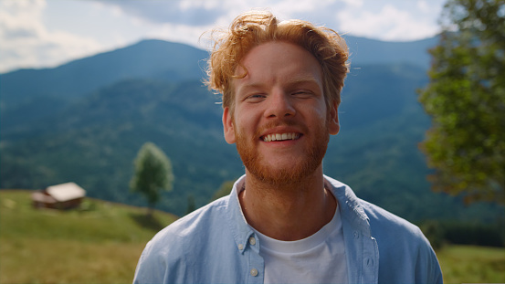Portrait of happy man smiling on standing green meadow sunny day. Positive red hair guy relaxing in summer vacation outdoors. Cheerful person looking camera in front beautiful mountains close up.