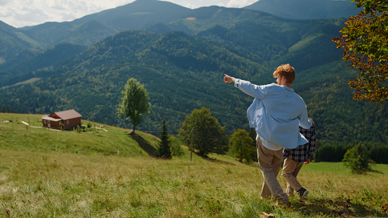 Back view of young father hugging son walking on mountain hill. Unknown redhair man going down green slope with cute boy. Carefree guy showing nature views to child outdoors. Family holiday concept.