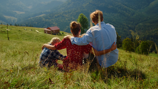 Unrecognizable family hugging sitting green grass hill. Unknown parents spending summer holiday with children in mountains. Back view of relaxed people enjoying beautiful sunny nature outdoors.