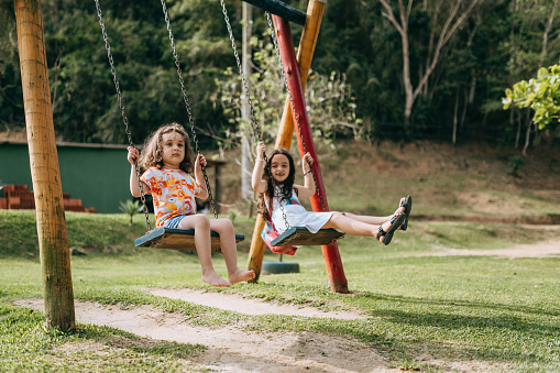 Mother and sister of liitle girls help to push the swing to make them fun and happy during holiday activity in playground of public park.