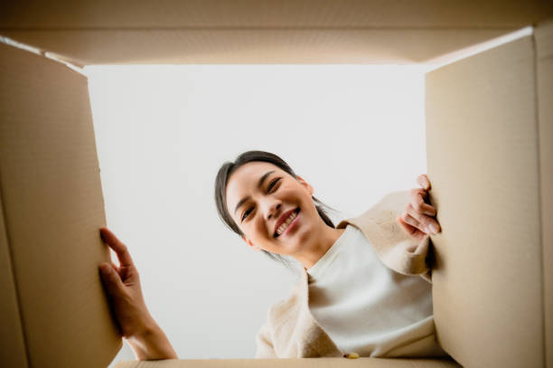 Surprised young asian woman unpacking. Opening carton box and looking inside. Packaging box, delivery service. Human emotions and facial expression stock photo
