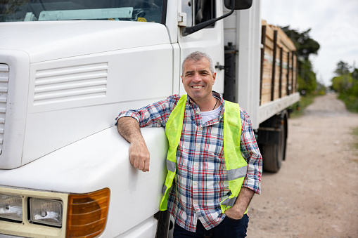 A warehouse driver is standing with arms crossed in front of the pickup. He is driving and shipping goods to all their clients. The client can be sure that the goods will come to them on time.