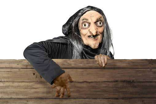 Old witch in a cloak standing behind a wooden board isolated over white background
