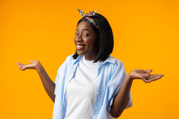 африканская молодая женщина, стоящая на желтом фоне с невежественным и растерянным выражением лица и поднятыми руками - blank expression head and shoulders horizontal studio shot стоковые фото и изображения