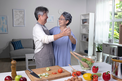 Beautiful Asian senior couple is dancing and smiling while cooking together in kitchen, Couple in love enjoying cooking healthy salad.