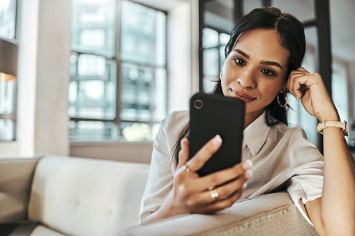 Woman, smartphone for social media and relax on living room sofa using app to scroll social network or watch online entertainment video. Indian woman, phone in hand and sitting on couch at home