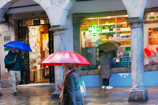 Santiago de Compostela, Spain- February 14, 2022: Green cross and commercial pharmacy sign , stone arches. Pedestrians walking in the street with umbrellas. Santiago de Compostela, Galicia, Spain.