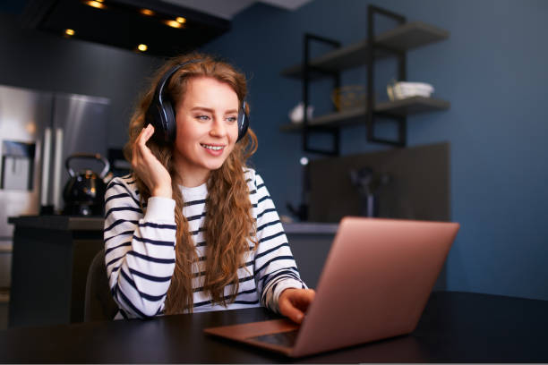 Customer support service call center manager speaking, working from home. Curly woman with headset, talks on video conference to laptop webcam. Young business woman consults business partners remotely stock photo