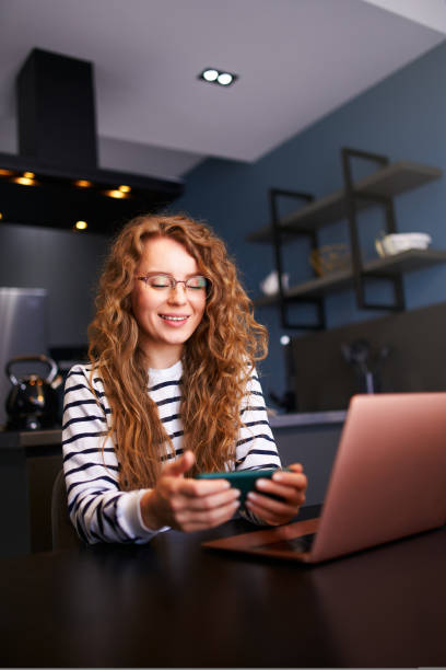 Pretty blonde woman distracted from work on the laptop watching video on smartphone. Freelancer holding mobile phone and browsing, playing mobile games and chatting with friends on remote work. stock photo