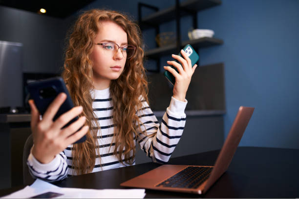 istock Curly blonde woman in glasses working from home with multiple electronic internet devices. Freelancer businesswoman has tablet, cellphone in hands and laptop on table. Multitasking theme. 1440453304