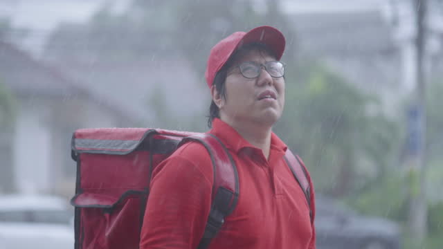 Delivery person taking off his cap and eyeglasses and wiping his face using arms while resting on the way to customer house in heavy rain.