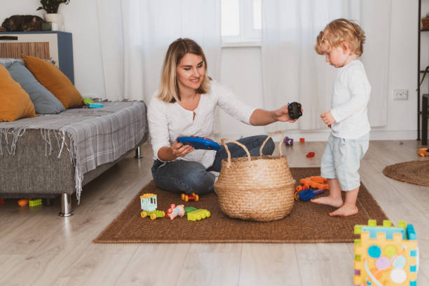 Young woman, housewife puts toys in the basket with her little son at home stock photo