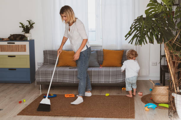 Young woman, housewife cleans up toys with broom together with her little son at home stock photo