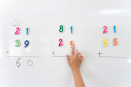 Primary school student in uniform solving math problem with magnetic numbers on whiteboard.