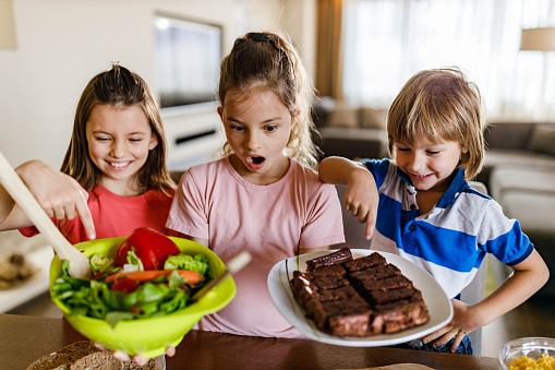 Little girl holding fresh salad and sweet cookies while her siblings choosing their favorite food.