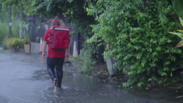 Rear view of delivery man wading through flooded road in the village while it's raining heavily.