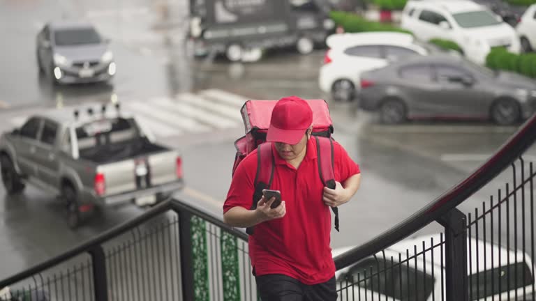 Delivery man walking up the stairs of footbridge, looking at his smart phone while searching for his destination on a rainy day.