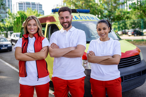 Multi-ethnic group of paramedics standing at the side of an ambulance with open doors. Their coworker carrying a medical trauma bag. They are smiling at the camera.