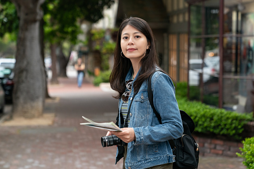 solo travel lifestyle and people in usa. portrait of smiling asian girl turning to look at camera with camera and guide map in hand on the street in Carmel by the sea California