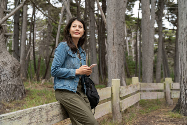 smiling asian chinese woman traveler sitting relaxing on low fence and enjoying natural beauty while visiting the crocker grove along 17 mile drive in california usa - lone cypress tree imagens e fotografias de stock