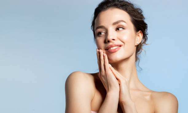 Young girl with hands near skin face Beautiful woman hold folded palm near face. Millennial lady enjoying soft smooth skin on hand face. Smiling model looking aside. Studio portrait on blue beauty product photography stock pictures, royalty-free photos & images