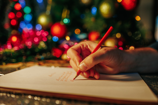 christmas time Blurred background gift wrapping and colorful tinsel unrecognizable human writing hand. Red pencil in hand. Writes a letter. Selective focus.