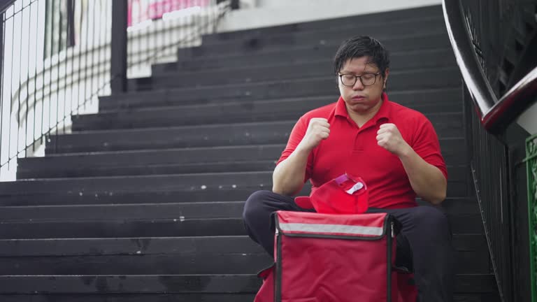 Delivery man sitting on a step of footbridge to rest, looking up and sighing. Then encouraging himself by raising his arms, standing up and walking to work.