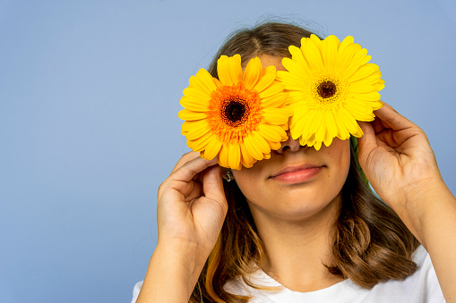 A happy pretty girl closes her eyes with yellow gerbera flowers. blue background. International Women's Day. A joyful girl with flowers in her eyes is fooling around. spring mood, positive attitude