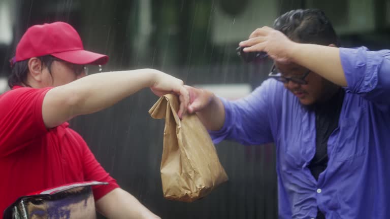 Delivery guy opening food delivery box and handing paper bag to customer while it's raining cats and dogs. Then customer walking back to his front door.