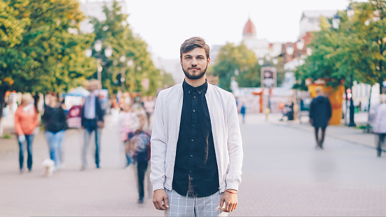 portrait of serious young man looking at camera standing in center of busy pedestrian street in summer wearing stylish clothing while many people are passing by.