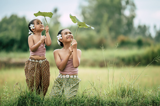 Portrait of lovely sister and young sister in Thai traditional dress and put white flower on her ear, looking to lotus' leaf in hand and smile with happy on rice field, copy space