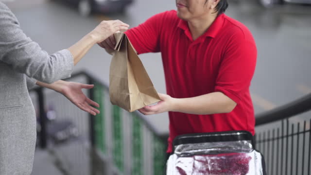 Delivery man opening food delivery box and handing a paper bag to businesswoman on footbridge and then carrying delivery box on shoulders and walking down.