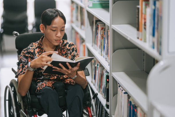 Asian teenage boy with disability in wheelchair reading book in library stock photo