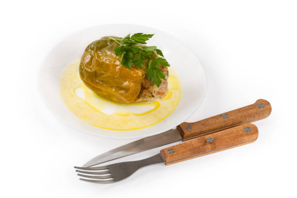Cooked green with yellow bell pepper stuffed with rice and chopped meat on the white dish, fork and knife on a white background