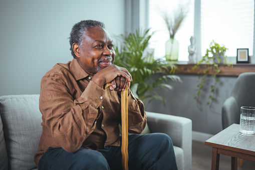 Thoughtful elderly man sitting alone at home with his walking cane. Shot of a senior man sitting at home. Indoor shot of happy elderly man sitting at home with walking stock and smiling