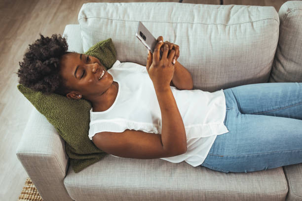 istock Happy black woman using smart phone and text messaging while relaxing at night in the living room. 1440482944