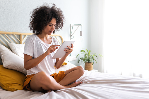 Young multiracial woman using sitting on bed down relaxing at home cozy bedroom. Copy space.Technology concept.