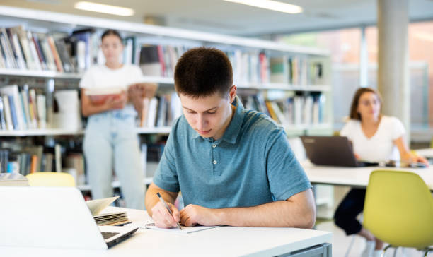 istock Concentrated young man checking plans in calendar while studying in the library and using laptop 1440489971