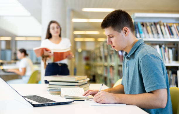 istock Focused casual young male writing in planner and using laptop in the library 1440489980