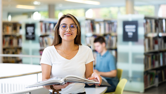 Happy Latin woman in casual clothes standing with open book in the middle of the library, looking at camera