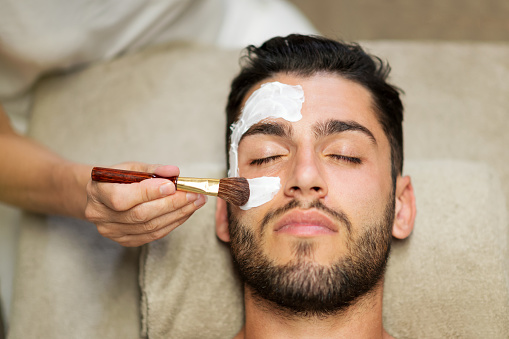 From above crop beautician applying clay mask on face of calm handsome bearded man lying on table in spa salon